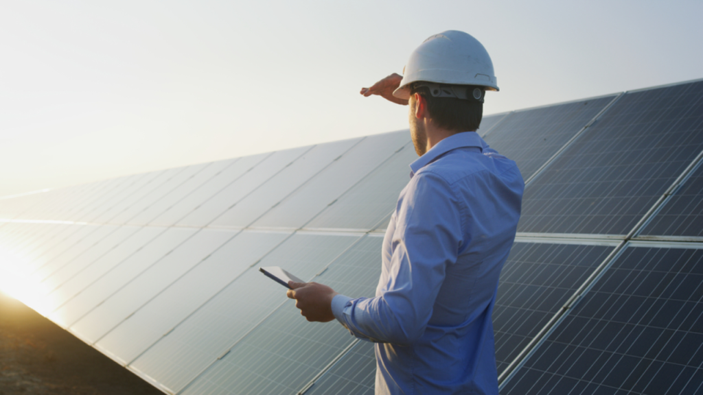 A imagem mostra um homem de capacete, camisa de cor clara, olhando em direção ao sol e a placas de energia fotovoltaica.